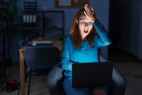 Brunette woman working at the office at night surprised with hand on head for mistake, remember error. forgot, bad memory concept.