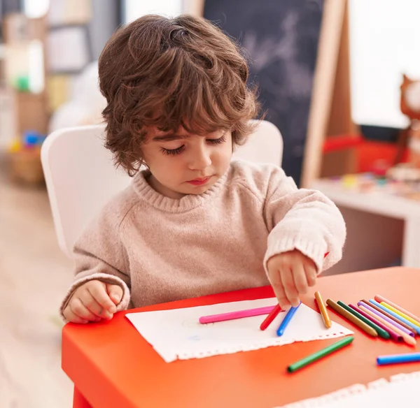 Adorable hispanic boy preschool student sitting on table drawing on paper at kindergarten