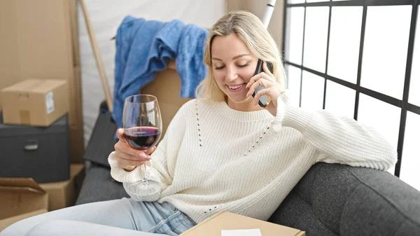 Young blonde woman talking on the smartphone and drinking wine at new home
