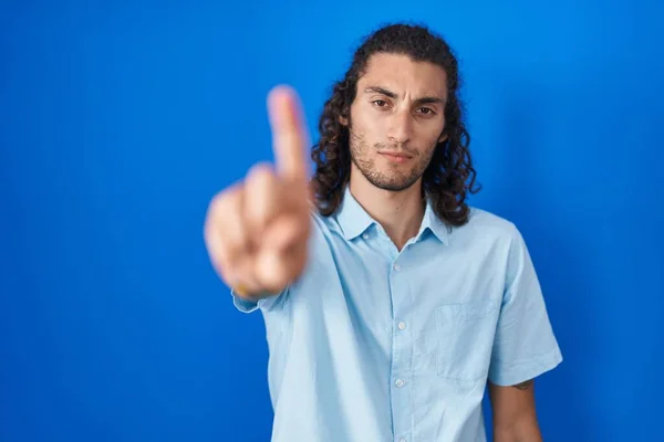 青い背景の上に立っている若いヒスパニック系の男は指を上げて怒りの表情で ジェスチャーを示していません — ストック写真
