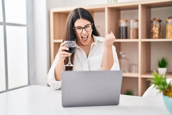 stock image Young brunette woman doing video call with laptop drinking a glass of wine screaming proud, celebrating victory and success very excited with raised arm 