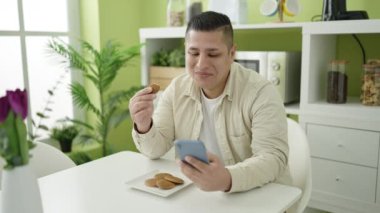 Genç İspanyol adam yemek odasında akıllı telefon kullanarak kurabiye yiyor.