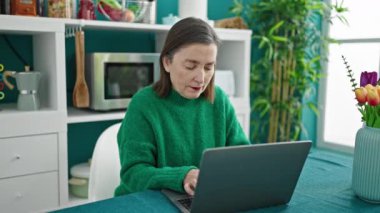 Yemek odasında gülümseyen dizüstü bilgisayar kullanan gri saçlı olgun İspanyol kadın.