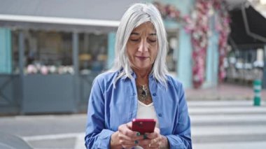 Orta yaşlı, kır saçlı, akıllı telefon kullanan, kahve dükkanının terasında gülümseyen bir kadın.