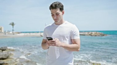 Genç İspanyol adam sahilde akıllı telefon kullanarak gülümsüyor.