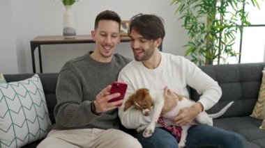 İki erkek akıllı telefondan selfie çekiyor. Kanepede köpeğiyle oturuyor.