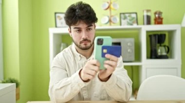 Genç İspanyol adam akıllı telefon ve kredi kartıyla alışveriş yapıyor yemek odasında masada oturuyor.