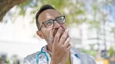 Orta yaşlı bir doktor şüpheli bir ifadeyle parkta durup düşünüyor.