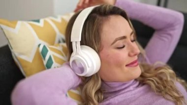 Genç sarışın kadın evdeki kanepede uzanmış müzik dinliyor.