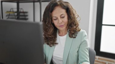 Orta yaşlı İspanyol iş kadını. Ofiste bilgisayar kullanıyor. Akıllı telefondan konuşuyor.