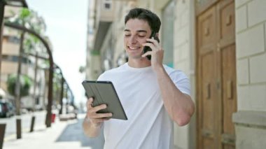 Young hispanic man talking on smartphone using touchpad at street