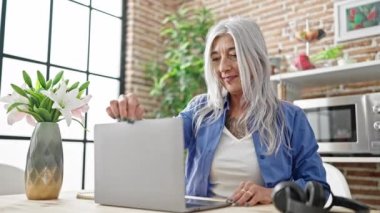 Orta yaşlı, gri saçlı bir kadın yemek odasındaki masada dizüstü bilgisayar kullanıyor.