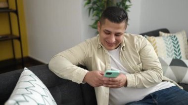 Genç İspanyol adam akıllı telefon kullanıyor. Kanepede oturuyor ve iyi bir jest yapıyor.