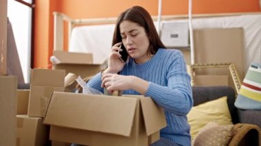 Genç beyaz kadın telefonda konuşuyor. Yeni evdeki karton kutuları açıyor.