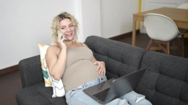 Akıllı telefondan konuşan hamile bir kadın dizüstü bilgisayarla evde karnına dokunuyor.