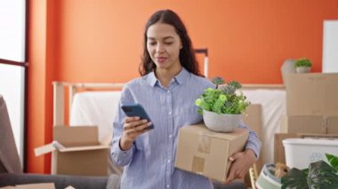 Genç, güzel, İspanyol bir kadın yeni evinde elinde bir kutu tutarak akıllı telefon kullanıyor.