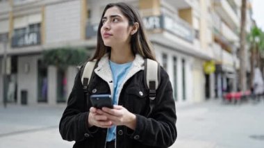 Genç, güzel, İspanyol bir kız öğrenci sokakta rahat bir ifadeyle akıllı telefon kullanıyor.