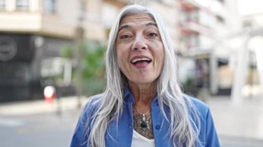 Orta yaşlı, kır saçlı bir kadın sokakta sürpriz bir ifadeyle duruyor.
