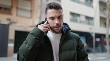 Young hispanic man talking on smartphone with serious expression at street