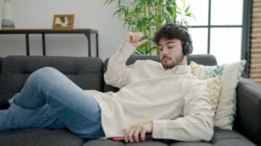 Genç İspanyol adam evde kanepede uzanmış akıllı telefonuyla müzik dinliyor.