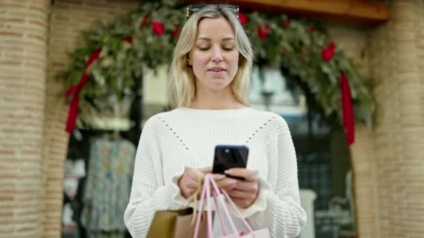 年轻的金发女人带着购物袋走在街上用智能手机 — 图库视频影像