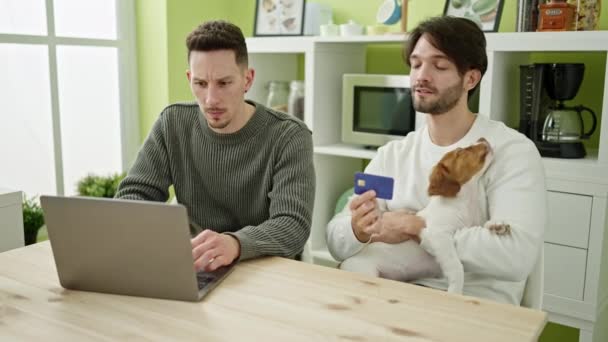 2人の男性カップルショッピングノートパソコンとクレジットカードでテーブルの上に座っている犬とダイニングルーム — ストック動画
