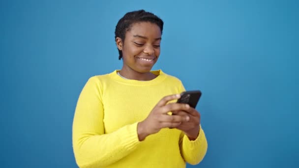 アフリカ系アメリカ人の女性はスマートフォンを使って孤立した青の背景に微笑む — ストック動画