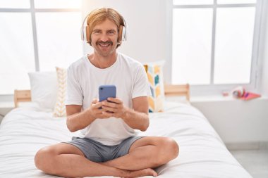 Young blond man listening to music sitting on bed at bedroom