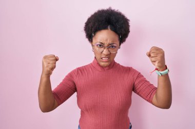 Pembe arka planda kıvırcık saçlı güzel bir Afrikalı kadın öfkeyle bağırırken kızgın ve kızgın yumruklarını kaldırıyor. öfke ve agresif konsept. 
