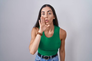 Young woman standing over isolated background hand on mouth telling secret rumor, whispering malicious talk conversation 