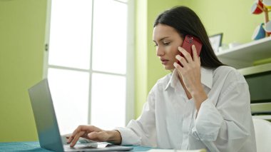 Genç ve güzel İspanyol kadın yemek odasında dizüstü bilgisayar kullanıyor ve akıllı telefondan konuşuyor.