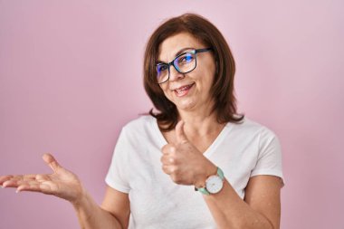 Middle age hispanic woman standing over pink background showing palm hand and doing ok gesture with thumbs up, smiling happy and cheerful 