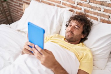 Young hispanic man watching video on touchpad lying on bed at bedroom