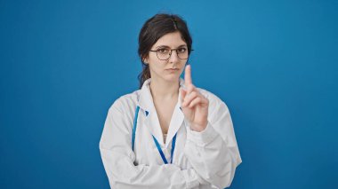 Genç, güzel İspanyol kadın doktor ciddi bir ifadeyle, izole edilmiş mavi arka planda parmağıyla hayır diyor.