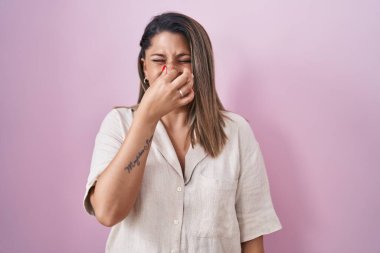 Blonde woman standing over pink background smelling something stinky and disgusting, intolerable smell, holding breath with fingers on nose. bad smell 