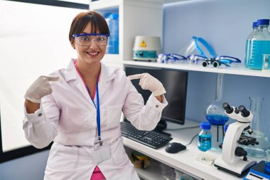 Young brunette woman working at scientist laboratory looking confident with smile on face, pointing oneself with fingers proud and happy. 