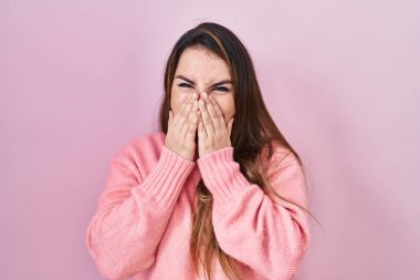 Young hispanic woman standing over pink background laughing and embarrassed giggle covering mouth with hands, gossip and scandal concept 