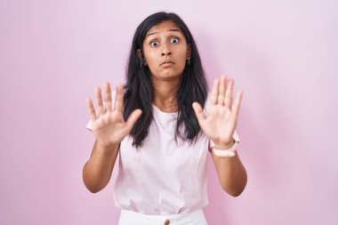 Young hispanic woman standing over pink background moving away hands palms showing refusal and denial with afraid and disgusting expression. stop and forbidden. 