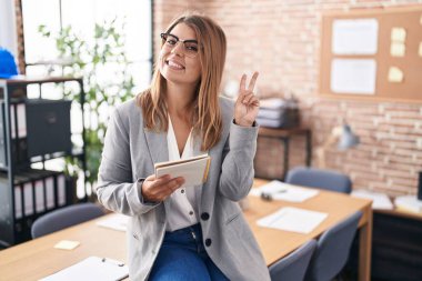 Ofiste çalışan, gözlüklü, gülümseyen, parmaklarını zafer işareti yaparken gösteren genç İspanyol kadın. İki numara.. 