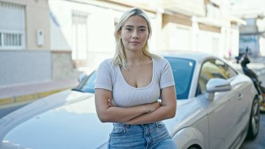 Genç, güzel, İspanyol bir kadın arabanın yanında duruyor. Ciddi bir ifade ve kollarını kavuşturmuş bir şekilde sokakta duruyor.