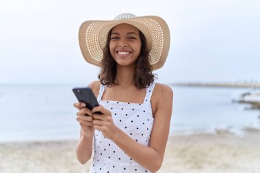 Young african american woman wearing summer hat using smartphone at seaside