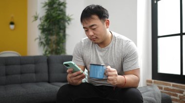 Genç Çinli adam akıllı telefon kullanıyor. Evde kahve içiyor.