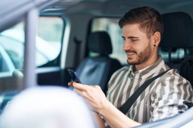 Beyaz bir adam akıllı telefon kullanıyor. Sokakta arabada oturuyor.