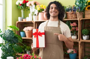 Çiçekçide çalışan kıvırcık saçlı İspanyol bir adam elinde hediyeyle mutlu bir şekilde elini ve parmağını göstererek gülümsüyor. 