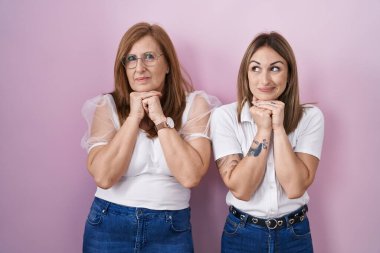 İspanyol anne ve kızı pembe arka planda günlük beyaz t-shirt giyiyorlar. Gergin ve heyecanlı bir şekilde çeneleri yana bakıyor. 