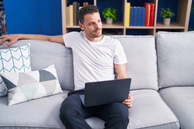 Young hispanic man using laptop sitting on sofa at home