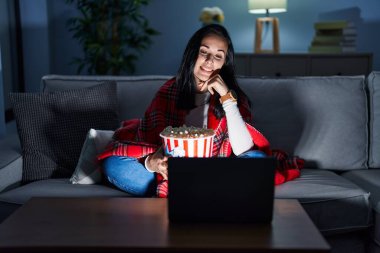 İspanyol kadın kanepede patlamış mısır yiyor. Elleri çenesinde, film seyrediyor. Soru sormayı düşünüyor. Gülümseyen ve düşünceli bir yüz. şüphe kavramı. 