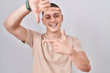 İzole edilmiş arka planda duran genç adam gülümsüyor ve mutlu yüzlü eller ve parmaklarla çerçeve yapıyor. yaratıcılık ve fotoğrafçılık kavramı. 