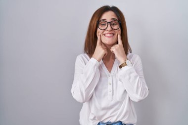Esmer kadın izole edilmiş beyaz arka planda duruyor ağzı açık gülümsüyor, parmakları işaret ediyor ve zorla neşeli bir gülümseme sergiliyor. 