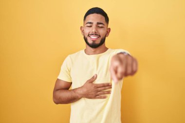 Young hispanic man standing over yellow background laughing at you, pointing finger to the camera with hand over body, shame expression 